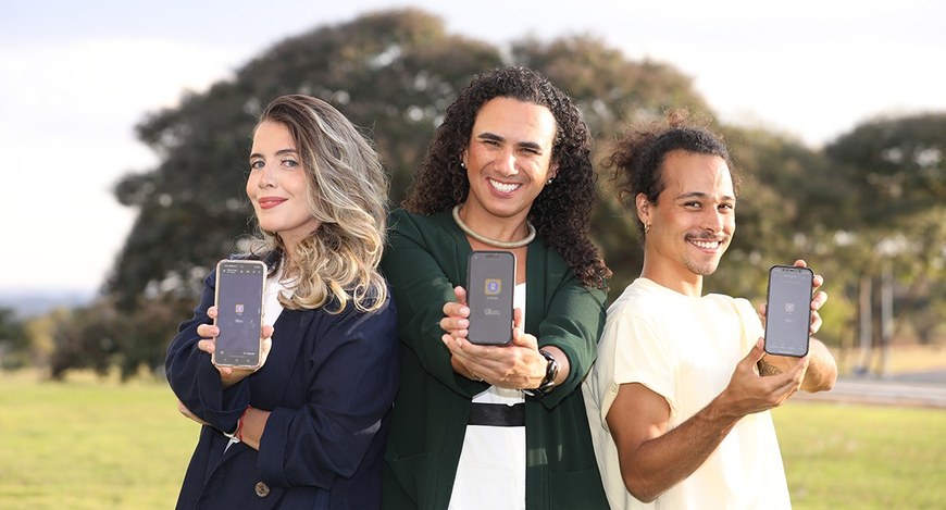 Três pessoas estão lado a lado, ao ar livre, sorrindo e segurando celulares voltados para a câme...