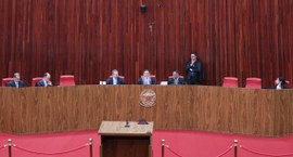 Dois homens vestindo togas pretas com detalhes vermelhos sobre ternos azuis caminham lado a lado...