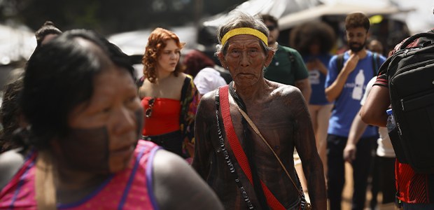 A imagem mostra um homem indígena no centro, com o corpo pintado de preto e usando uma faixa ver...