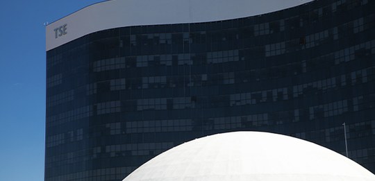 Edifício do Tribunal Superior Eleitoral (TSE), com uma fachada moderna de vidro escuro. No topo ...