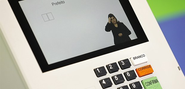 A imagem mostra uma urna eletrônica utilizada em eleições no Brasil. A tela da urna exibe a pala...