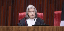 A imagem mostra a ministra Carmen Lúcia sentada em frente a uma bancada de madeira em um ambient...