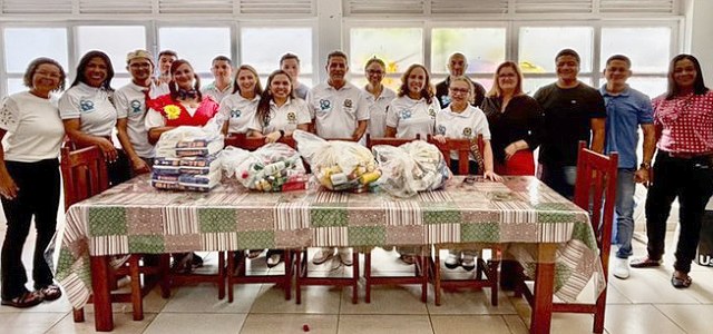 Fotografia colorida mostra um grupo de cerca de 20 pessoas em pé, lado a lado, atrás de uma mesa...