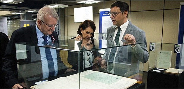 Essa foto mostra três pessoas, duas olhando atenciosamente para o interior de uma vitrine de vid...