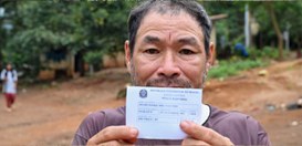 A foto mostra um homem em primeiro plano, olhando diretamente para a câmera, segurando um docume...