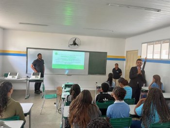 A imagem mostra um grupo de estudantes e adultos em uma sala de aula, participando de uma ativid...