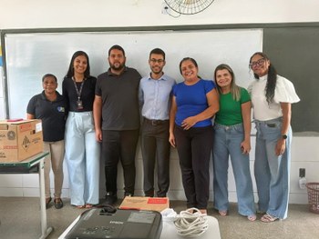 A imagem mostra um grupo de estudantes e adultos em uma sala de aula, participando de uma ativid...
