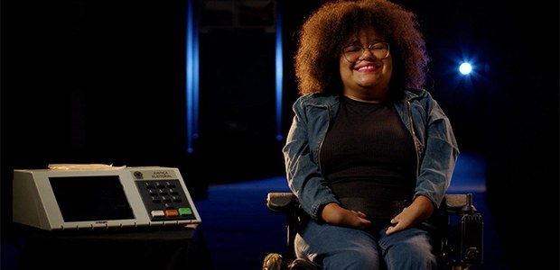 Jovem cadeirante ao lado da urna eletrônica, que dispõe de diversos recursos de acessibilidade - 21.09.2025 Mulher negra ,jovem e cadeirante ao lado da urna eletrônica de votação