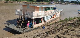 Fotografia mostra uma embarcação de madeira parada na margem de um rio de água barrenta. O barco...