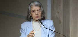 A imagem mostra a ministra Cármen Lúcia falando em um microfone durante um evento. Ela tem cabel...