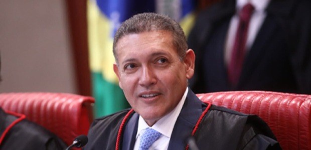 A imagem mostra  o presidente do TSE, Nunes Marques. Ele está sentado em uma cadeira vermelha, v...
