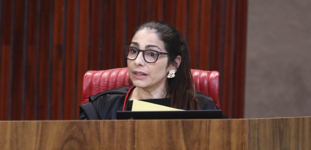 A imagem mostra a ministra Estela aranha sentada em uma cadeira alta e estofada na cor vermelha,...