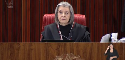 A imagem mostra a Ministra Cármen Lúcia sentada em uma cadeira vermelha, vestindo uma toga preta...