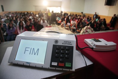 Urna eletrônica sobre uma mesa, à frente de um auditório com muitas pessoas.