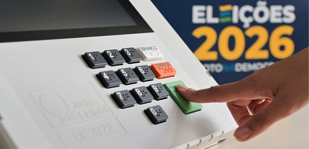Mão pressiona a tecla Confirma de uma urna eletrônica, com identificação das Eleições 2026 ao fundo.