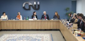 Fotografia de uma sala de reuniões onde diversas pessoas formalmente vestidas estão dispostas ao...