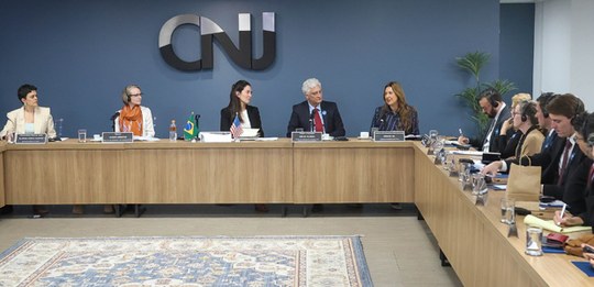 Fotografia de uma sala de reuniões onde diversas pessoas formalmente vestidas estão dispostas ao...